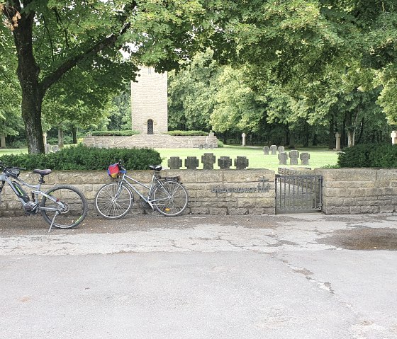 Twee fietsen leunen tegen een stenen muur. Achter hen ligt een begraafplaats met grafstenen en een toren omringd door bomen., © TI Bitburger Land