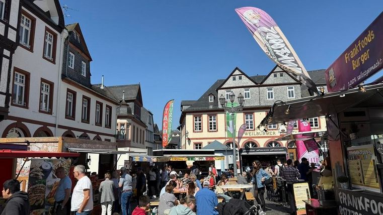 A bustling market with many people and food stalls. In the background, picturesque old buildings and a clear blue sky can be seen.