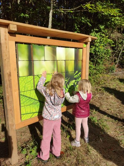 Zwei Kinder erkunden eine Lernstation im Wald mit einem großen Holzrahmen und grünen Tafeln.