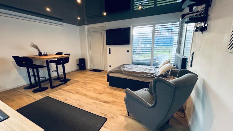 A modern room with a dining table, a comfortable armchair, and a large window. The decor is bright and inviting.