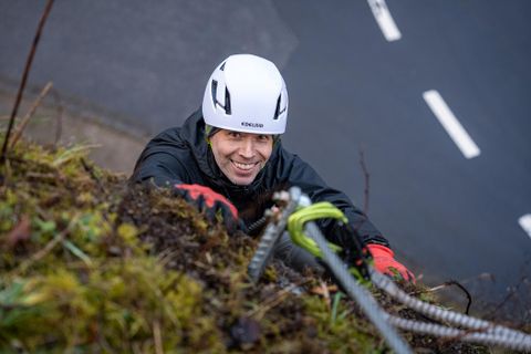 A climber with a helmet is smiling while climbing up a steep rock face. The surroundings are green, and the path is visible in the background.