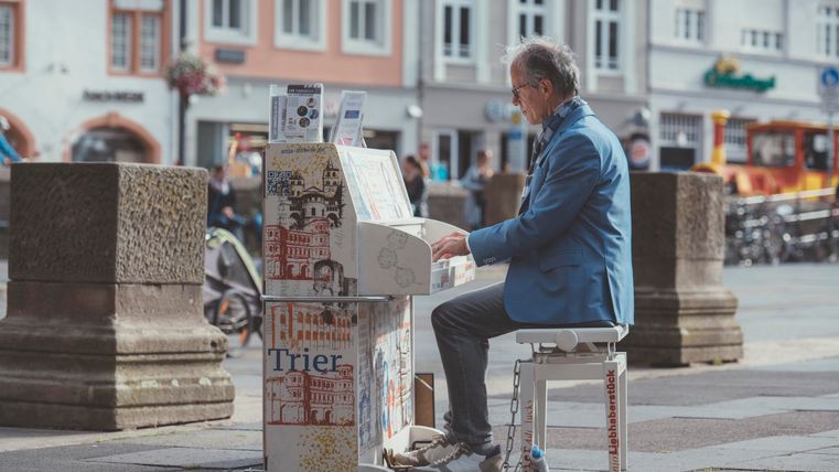 Ein Mann spielt Klavier auf einem belebten Platz. Im Hintergrund sind historische Gebäude und Fahrräder zu sehen.