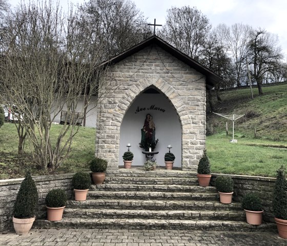 Petite chapelle en pierre avec une statue de la Vierge Marie, entourée de plantes en pots. La chapelle se trouve sur un escalier, avec des arbres et une prairie en arrière-plan., © TI Bitburger Land - Biggi Thiebach