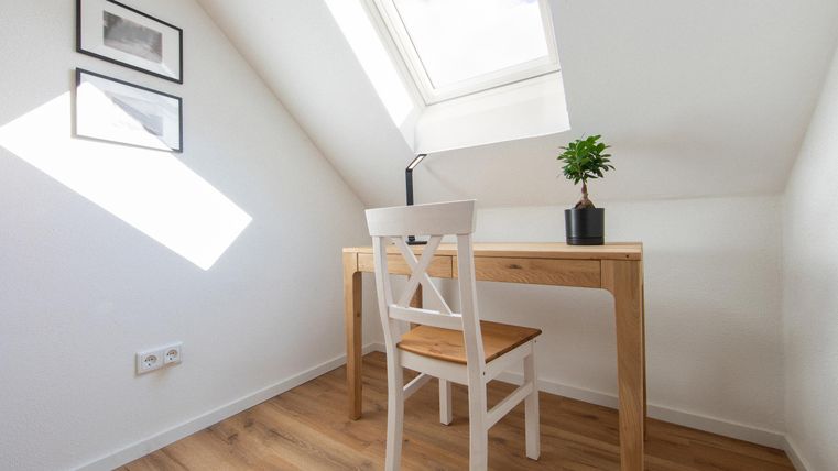 Ein helles Arbeitszimmer mit einem Schreibtisch, einer weißen Stuhl und einer kleinen Pflanze. Große Fenster sorgen für viel Tageslicht.