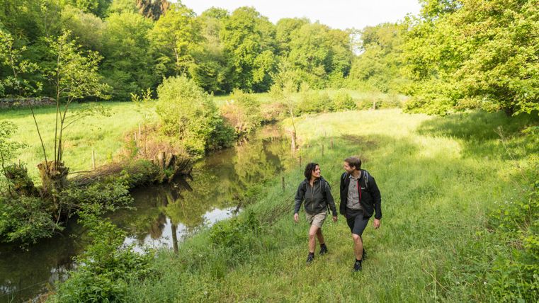 Twee wandelaars lopen langs een rivier in een groen landschap.