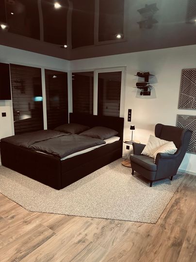 Une chambre moderne avec un grand lit double et un fauteuil confortable. La pièce a un parquet en bois chaleureux et de grandes fenêtres qui laissent entrer beaucoup de lumière.