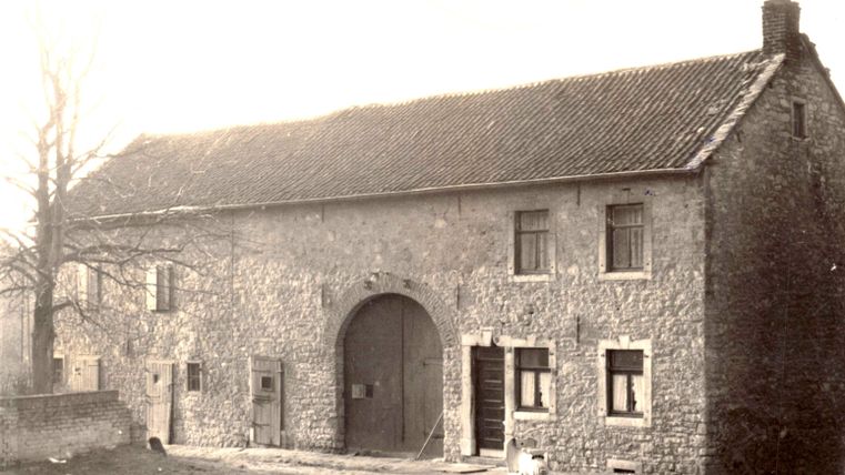 Une vieille maison en pierre dotée d'un grand portail et de plusieurs fenêtres. Au premier plan, on aperçoit quelques arbres et une route dégagée.