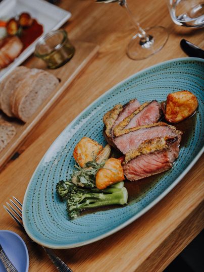 Een kunstzinnig opgemaakt bord met mals vlees, aardappelen en broccoli. Op de achtergrond zijn brood en een glas wijn te zien.