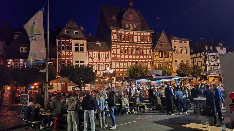 Ein belebter Marktplatz bei Nacht mit historischen, Fachwerkhäusern. Menschen stehen zusammen, während Stände Essen und Getränke anbieten.