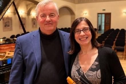Zwei Personen stehen in einem Konzertsaal, in dem ein Klavier sichtbar ist. Der Mann trägt einen blauen Anzug, die Frau hält ein Instrument und lächelt.
