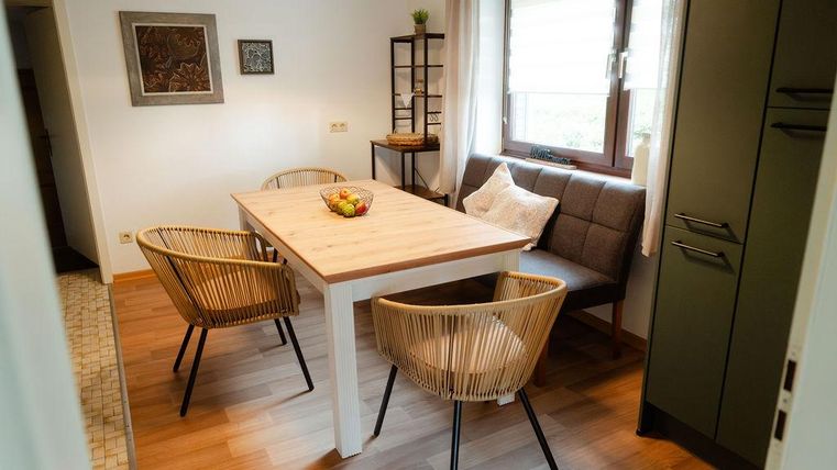 Une salle à manger confortable avec une table en bois et des chaises. La lumière naturelle entre par la fenêtre, et des fruits frais sont sur la table.