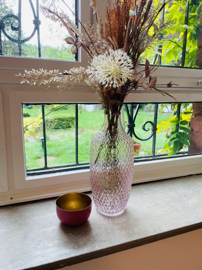 Un vase avec des fleurs séchées est posée sur un appui de fenêtre. À côté de la vase se trouve un petit bol coloré.