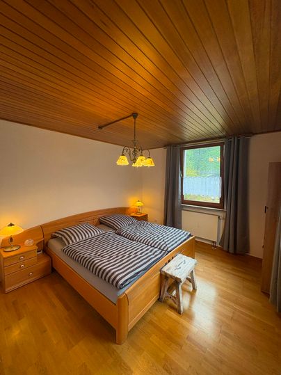 A cozy bedroom with a double bed and wooden ceiling. There are nightstands, a floor lamp, and a window with curtains.