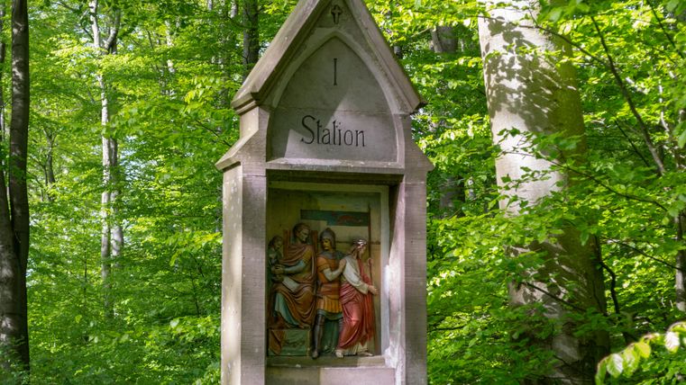 Un pilier de pierre dans la forêt, portant l'inscription 'I Station', montre une scène religieuse.