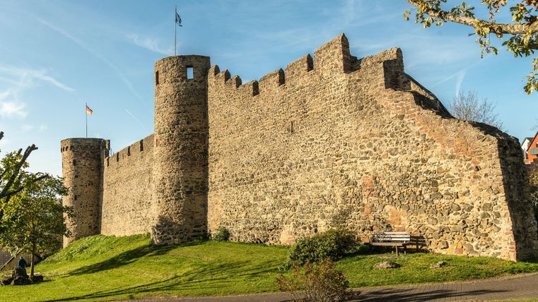 Eine alte Burg mit starken Mauern und Türmen. Grüne Wiesen umgeben die Festung und der Himmel ist blau.