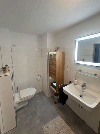 A modern bathroom with bright tiles and a spacious shower. There is a sink, a toilet, and a wooden cabinet for additional storage space.