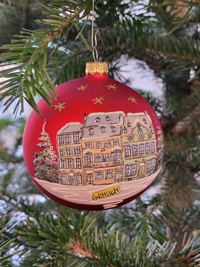 Er hangt een rode kerstbal aan een boom. Er staat een versierd gebouw en gouden sterren op afgebeeld.