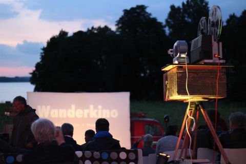 Eine Projektion mit dem Wort "Wanderkino" auf einer Leinwand vor einer Zuschauergruppe im Freien. Der Rahmen ist mit Bäumen und einem See im Hintergrund umgeben.