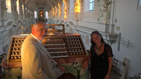 Two people are standing at a large, historical organ instrument. The room is brightly illuminated and has impressive architecture.