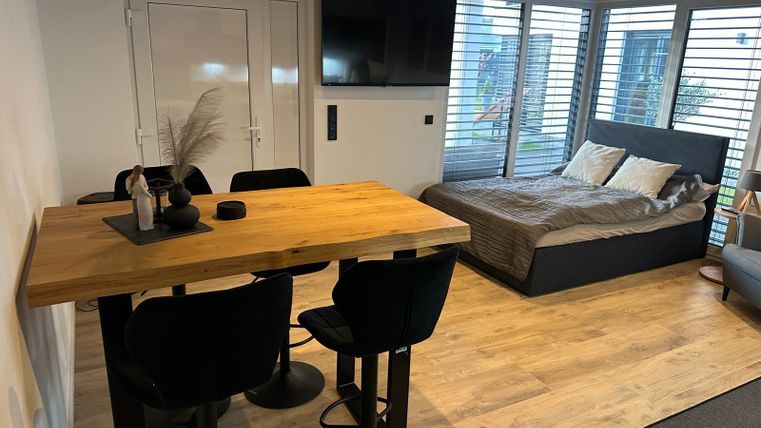 A modern living room with a dining table and chairs. In the background, there is a sleeping area with a cozy bed.