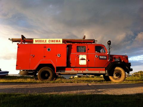 A red truck that serves as a mobile cinema is parked on a street. The clouds in the background create a dramatic atmosphere for the scene.