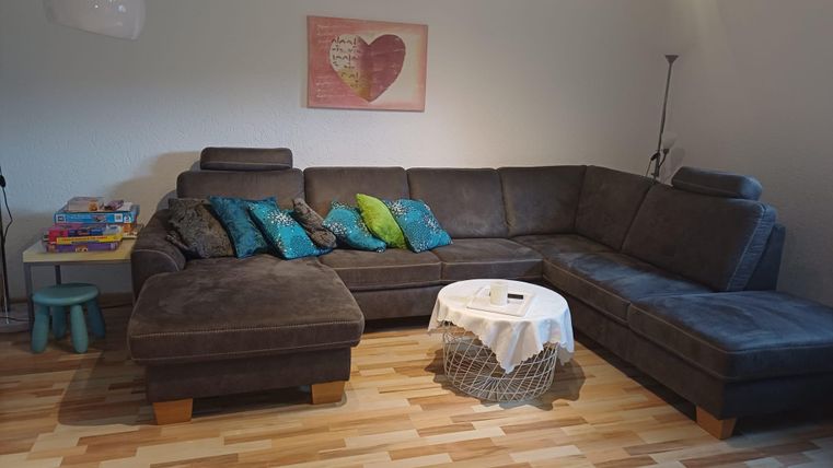 Un salon chaleureux avec un grand canapé gris et des coussins colorés. À côté, il y a une table avec une nappe et des livres sur une petite table.