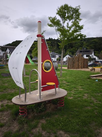 Een speeltoestel in de vorm van een boot met een wit zeil en een rood vlak. Op de achtergrond zijn bomen en andere speeltoestellen te zien.