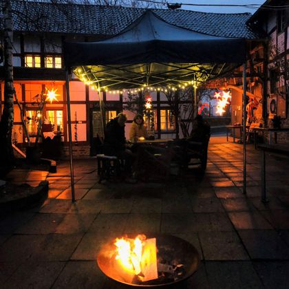 Eine gemütliche Terrasse mit einem Zeltdach, unter dem sich Personen treffen. Im Vordergrund brennt ein Feuer in einem metallenen Behälter, während die Umgebung in warmen Lichtern erleuchtet ist.