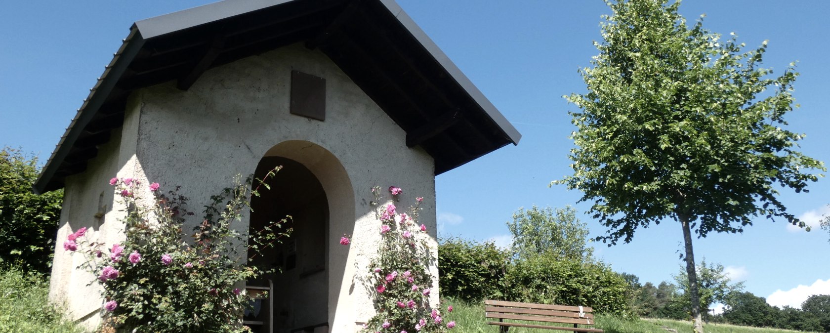 Kleine Kapelle im Grünen, umgeben von blühenden Rosen. Eine Bank steht daneben, ein Baum spendet Schatten. Blauer Himmel im Hintergrund., © Tourist-Information Bitburger Land, Melanie Salzburger