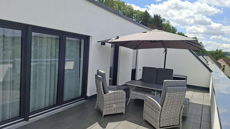 Eine moderne Terrasse mit einem Esstisch und bequemen Stühlen. Ein Sonnenschirm bietet Schatten, während die Natur im Hintergrund sichtbar ist.