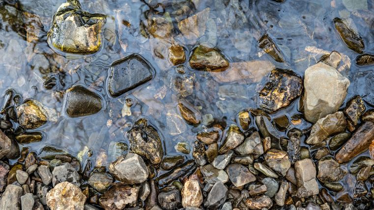 Een heldere stroom water die over ronde stenen stroomt. De verschillende stenen hebben verschillende kleuren en texturen.