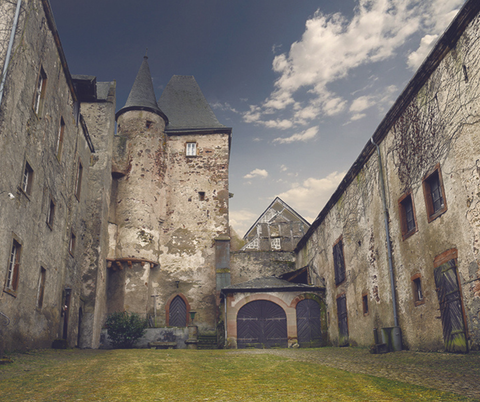 Une ancienne cour de château crépie avec une tour ronde à l'arrière-plan. La cour est entourée de bâtiments en pierre couverts de lierre.