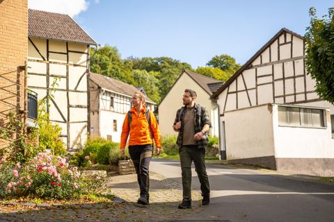 A pair of walkers strolls through a picturesque, quiet street with traditional half-timbered houses. The surroundings are green and friendly in the light of a sunny day.