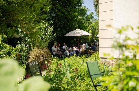 A sunny garden with guests sitting under sun umbrellas. Surrounded by many plants and an inviting atmosphere.
