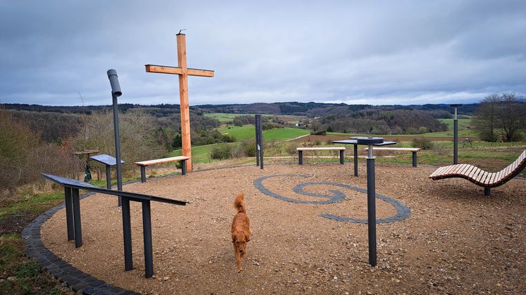 Ein Aussichtspunkt mit einer Holzskulptur in Form eines Kreuzes. Im Vordergrund läuft ein Hund über den Platz mit Blick auf die grünen Hügel.