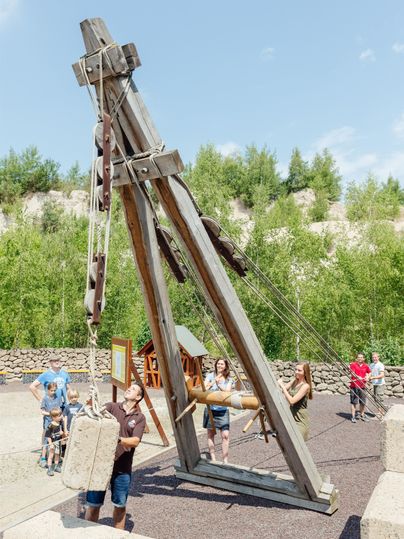 Eine große Holzmaschine steht auf einem Spielplatz. Kinder und Erwachsene interagieren mit der Maschine bei schönem Wetter.