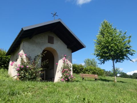 Petite chapelle dans la verdure avec des roses en fleurs et un banc à côté.
