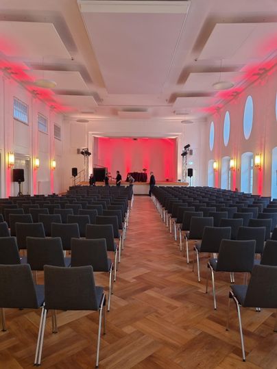Ein Veranstaltungsraum mit vielen Stühlen in Reihen. Die Wände sind mit rotem Licht beleuchtet und eine Bühne ist im Hintergrund sichtbar.