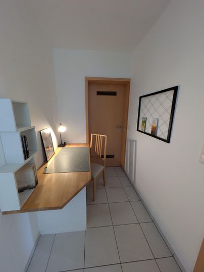 Een lichte gang met een bureau en een stoel. Aan de muur hangen een paar schilderijen, en er staat een lamp op de tafel.