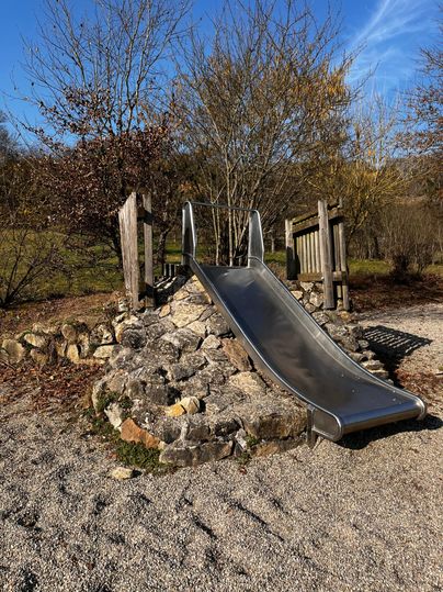 Een glijbaan van roestvrij staal op een speelplaats, omgeven door stenen en grind. Op de achtergrond zijn bomen en een blauwe lucht zichtbaar.