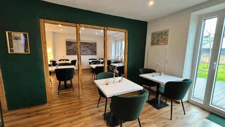 A modern restaurant with wooden floors and elegant green chairs. The tables are nicely set and the room has plenty of daylight.
