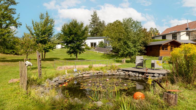 Een mooie tuin met een kleine vijver en bomen. Op de achtergrond zijn een huis en een tuinhuisje te zien.