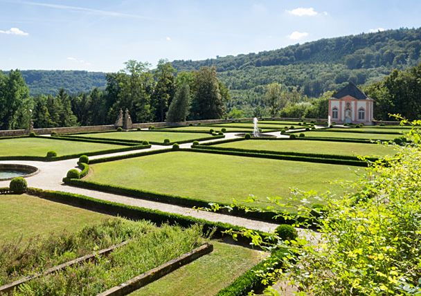 Jardin à la française avec des haies géométriques et des pelouses, au fond un petit bâtiment et des collines boisées.