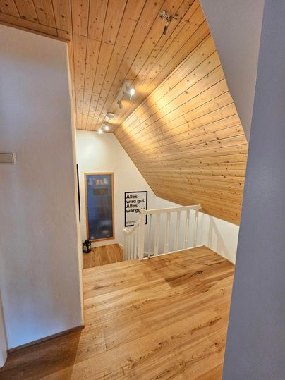 Een lichte gang met houten plafond en houten vloer. Een trap leidt naar een klein gebied met een raam en een kunstwerk aan de muur.