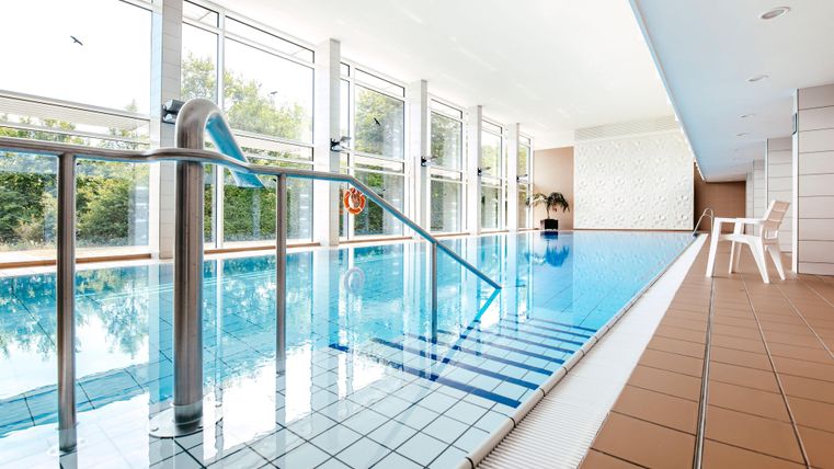 Ein moderner Innenpool mit glattem Wasser und großen Fenstern. Rechts steht ein weißer Stuhl und im Hintergrund sind Bäume sichtbar.