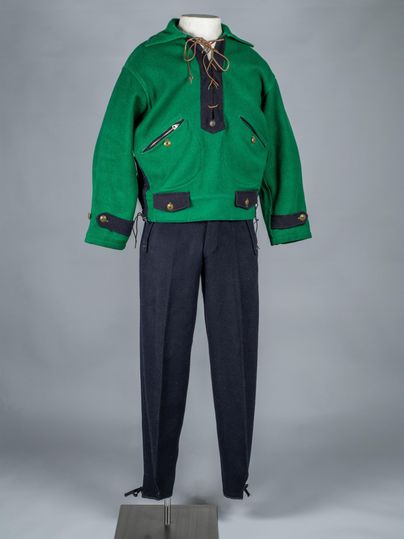 Een groene sweater met koord en twee voorzakken, evenals een donkere broek, zijn tentoongesteld op een mannequin. De kleding heeft een traditionele, sportieve uitstraling.