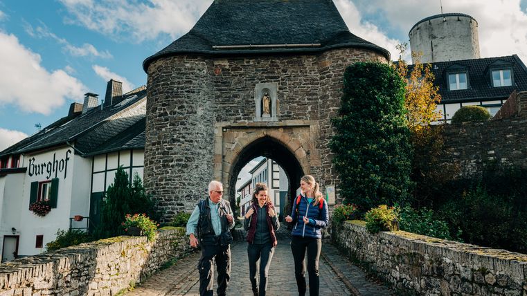 Trois personnes marchent sur un chemin pavé et franchissent une porte historique à Reifferscheid.
