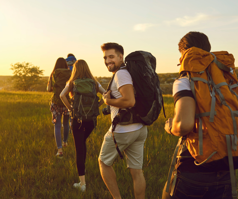 A group of young hikers with backpacks is walking across a green field. The sun is shining, creating a warm, inviting atmosphere.