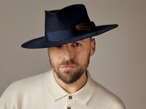 Een groep van zes muzikanten poseert in een gezellige ruimte. Ze staan en zitten rondom een rode fauteuil en dragen casual kleding.