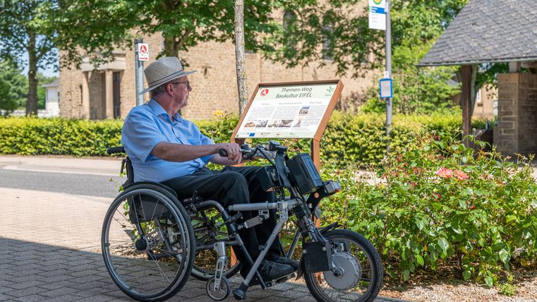 Een man in een rolstoel kijkt naar een bord voor het Eifel bouwcultuur themapad in Wolsfeld.